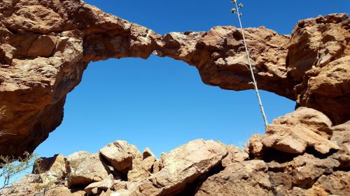 Natural rock arch against a clear blue sky, surrounded by rugged terrain and sparse vegetation.