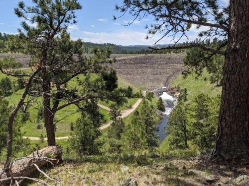 A scenic view of a river flowing past a dam, surrounded by green trees and hills under a clear blue sky.