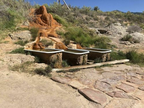 Three bathtubs sit on a grassy area near a natural hot spring, surrounded by rocky terrain and sparse vegetation.