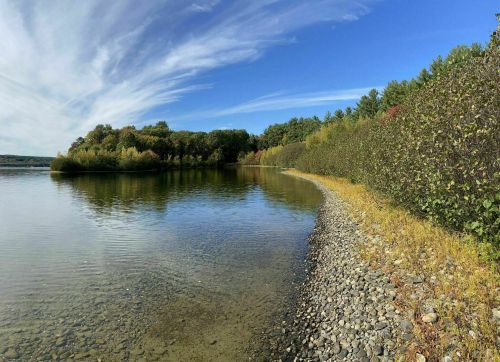 A serene lakeside view with clear water, rocky shore, and trees reflecting autumn colors under a blue sky.