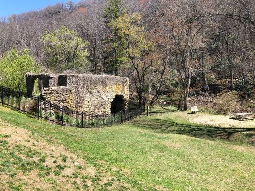Ruins of a stone structure surrounded by trees and grass, with a picnic area nearby in a wooded landscape.