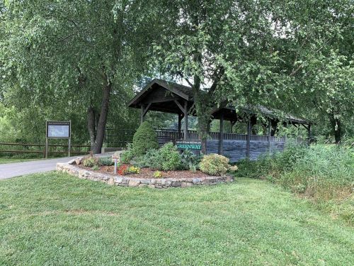 A wooden pavilion surrounded by greenery, with a sign for the Greenway and a landscaped flower bed in front.
