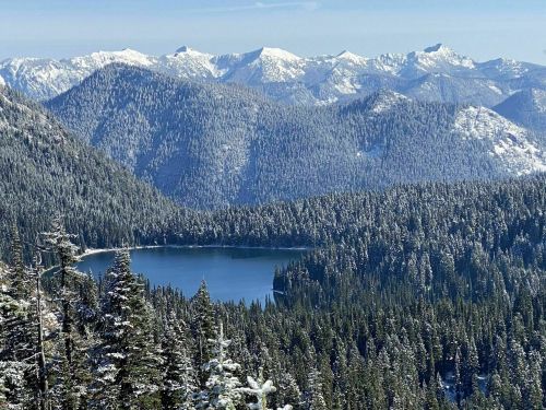 A serene mountain landscape featuring a snow-covered lake surrounded by evergreen trees and distant snow-capped peaks.