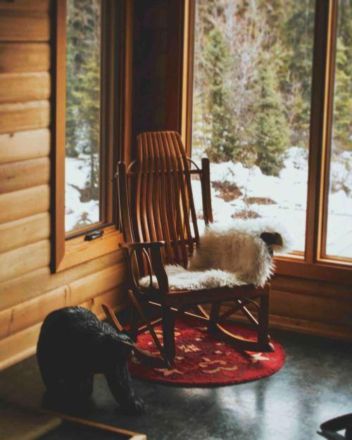 A cozy wooden rocking chair with a fur throw, beside a black bear figurine, in a cabin with snowy views outside.
