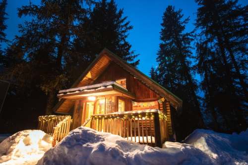 A cozy wooden cabin illuminated at night, surrounded by snow and tall evergreen trees.