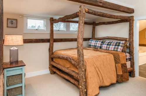 Cozy bedroom featuring a rustic log bed, plaid pillows, and a lamp beside a blue nightstand. Natural light from windows.