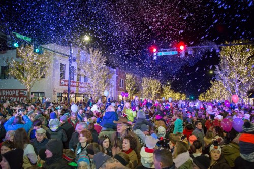 A festive crowd gathers on a snowy street, illuminated by holiday lights and decorations, celebrating together.