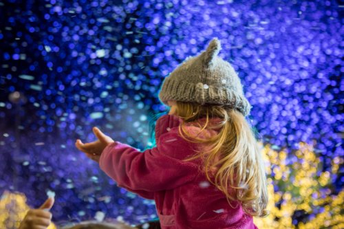 A young girl in a cozy hat and pink jacket joyfully reaches out as snowflakes fall around her.