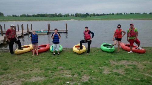 A group of six people in life jackets stand by colorful kayaks near a lake, ready for a kayaking adventure.