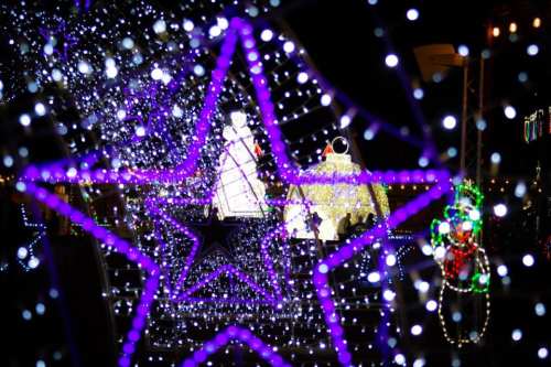 A vibrant display of purple star-shaped lights framing a festive scene with illuminated decorations in the background.