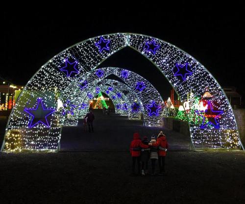 Three children in red jackets stand under illuminated arches decorated with blue stars and colorful holiday lights at night.