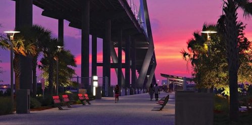 A scenic walkway under a bridge at sunset, with palm trees, benches, and vibrant purple and orange skies.