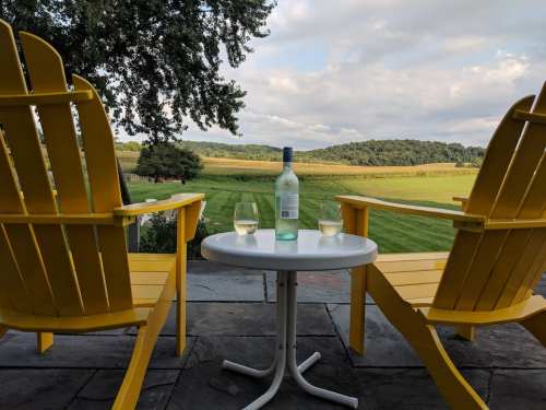 Two yellow chairs and a small table with wine glasses, overlooking a green field and distant hills under a cloudy sky.