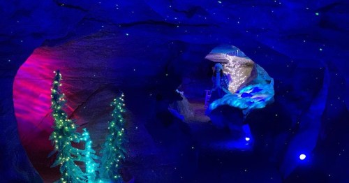 A colorful cave illuminated with blue lights, featuring decorated trees and a sparkling winter scene in the background.