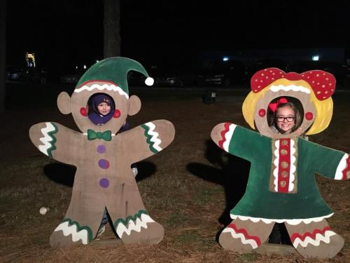 Two children peek through cutouts of gingerbread figures, one dressed as an elf and the other as a gingerbread girl, at night.