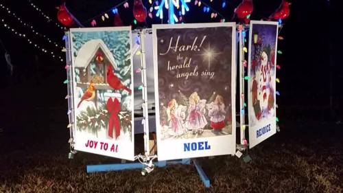 A festive display with three signs: "Joy to All," "Noel," and "Hark! The Herald Angels Sing," surrounded by colorful lights.
