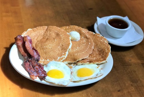 A plate of pancakes with butter, two fried eggs, crispy bacon, and a small bowl of syrup on the side.