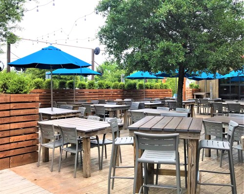 Outdoor dining area with wooden tables, gray chairs, and blue umbrellas under a tree, surrounded by greenery.
