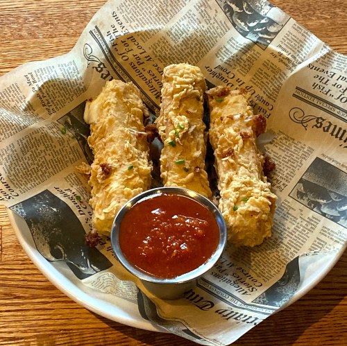 Three crispy fried items served on newspaper-style wrapping, accompanied by a small bowl of marinara sauce.