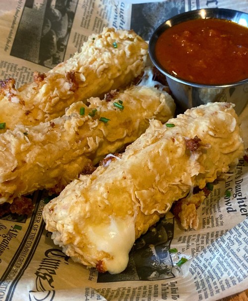 Three crispy fried cheese sticks served with a side of marinara sauce, placed on a bed of newspaper.
