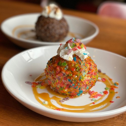 A colorful dessert ball coated in sprinkles, topped with whipped cream and drizzled with caramel sauce on a white plate.