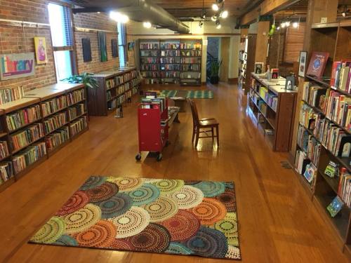 A cozy library with wooden floors, bookshelves, a colorful rug, and a book cart in a well-lit space.