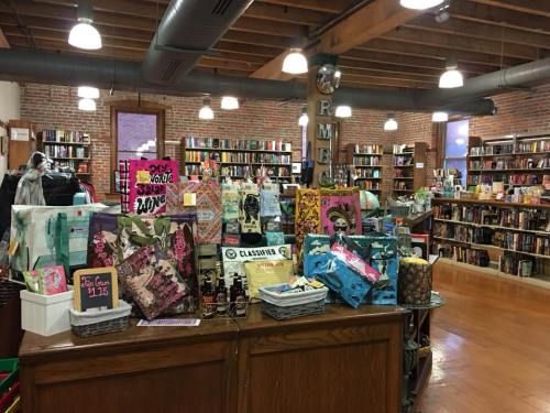 A cozy bookstore interior with shelves of books, a wooden counter displaying colorful bags and items.