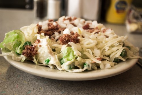 A plate of salad with lettuce, peas, bacon bits, and shredded cheese on a gray table.