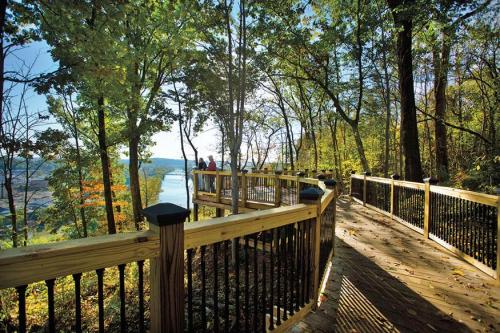 A wooden walkway winds through trees, overlooking a river with people enjoying the scenic view in the background.