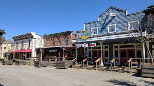 A sunny street scene featuring rustic storefronts with wooden facades and a clear blue sky above.