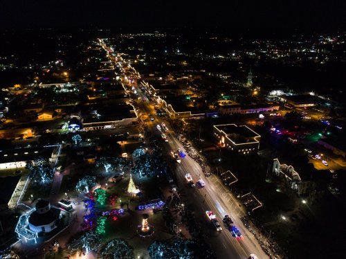Aerial view of a festive night parade with colorful lights lining the streets and a decorated park in the foreground.