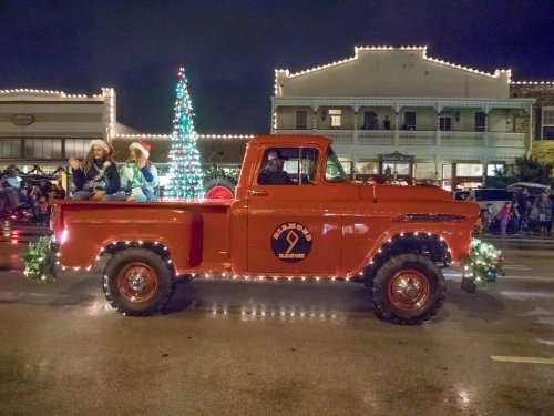 A festive red truck adorned with lights, carrying two people in holiday attire during a nighttime parade.