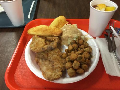 A red tray holds a plate with fried fish, okra, cornbread, and a side of creamy dish, alongside a cup of fruit.