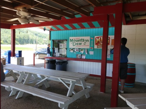 A colorful outdoor refreshment stand with picnic tables, featuring a Mountain Dew sign and a scenic view in the background.