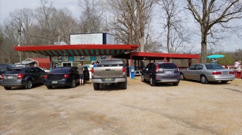 A colorful roadside eatery with cars parked outside, surrounded by trees and people enjoying the outdoor space.