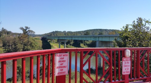 A red bridge overlooks a river with a highway bridge in the background and a warning sign about safety.