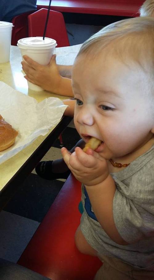 A toddler sitting at a table, eating a piece of food with a focused expression, surrounded by drinks and a hot dog.
