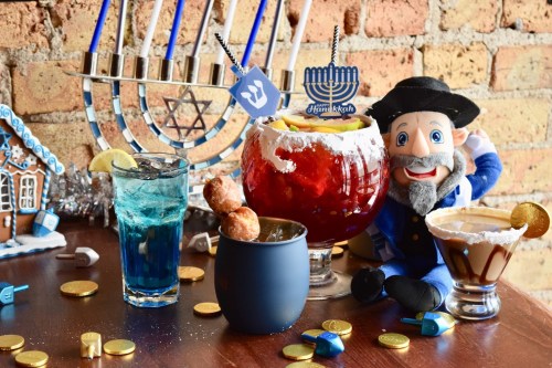 A festive table with colorful drinks, a plush character, and Hanukkah decorations, including a menorah and dreidels.