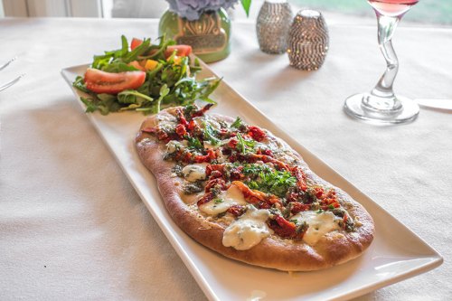 A flatbread topped with cheese, sun-dried tomatoes, and herbs, served with a side salad and a cocktail.