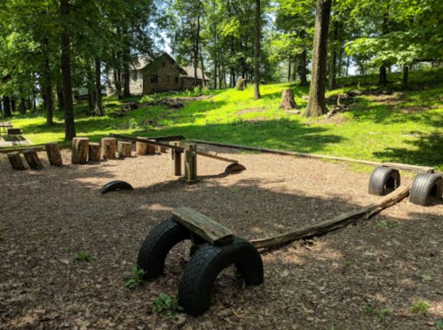 A natural playground with wooden structures and tire elements, surrounded by trees and a grassy hillside.