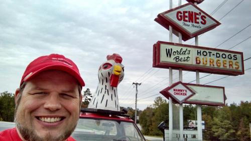 A smiling man poses with a chicken statue on his car, in front of a sign for Gene's Fine Foods and Mammy's Chicken.