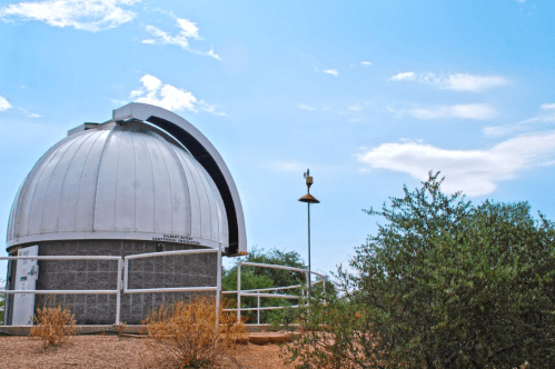 A white observatory dome against a blue sky, surrounded by sparse vegetation and a small weather station.