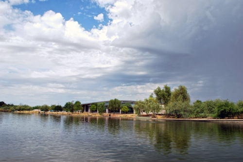 A serene lake scene with trees and a building under a partly cloudy sky. Reflections shimmer on the water's surface.