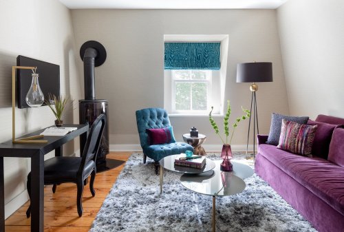 Cozy living room with a purple sofa, blue chair, glass coffee table, and a desk, featuring modern decor and natural light.