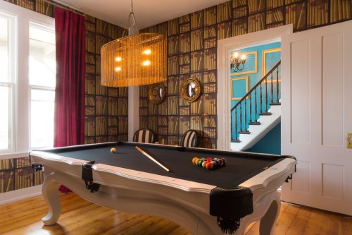A stylish game room with a white pool table, book-patterned wallpaper, and a staircase in the background.