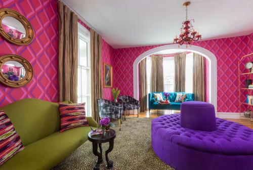 A vibrant living room with pink patterned walls, colorful furniture, and a plush purple ottoman.