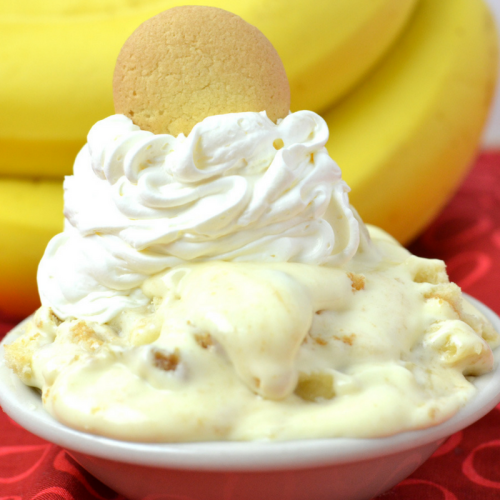 A bowl of banana pudding topped with whipped cream and a vanilla wafer, with bananas in the background.