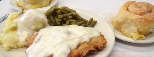 A plate of fried chicken with gravy, green beans, and mashed potatoes, alongside a cinnamon roll.
