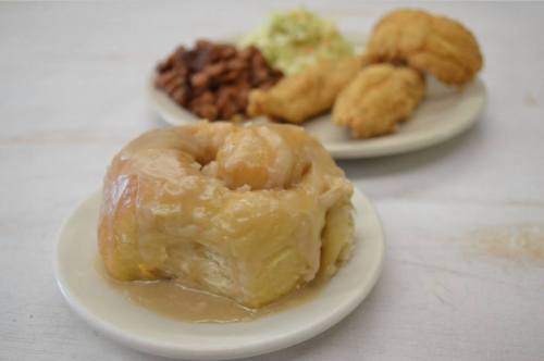 A plate featuring a glazed cinnamon roll, coleslaw, fried chicken, and baked beans.