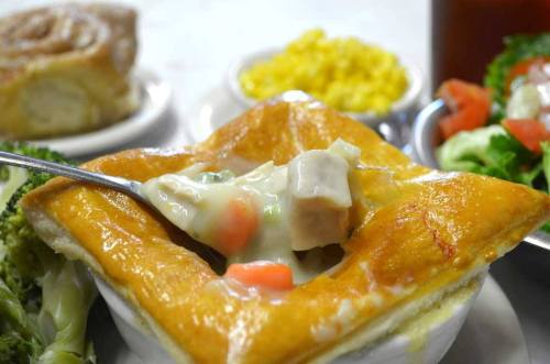 A bowl of chicken pot pie with a flaky crust, surrounded by broccoli, salad, and a side of yellow corn.
